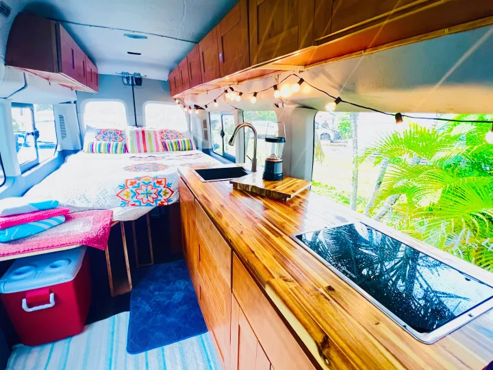 Explore this RV rental camper van interior: colorful bed, wooden kitchen countertop with sink & stove, and string lights.. Ford Transit Custom 2016