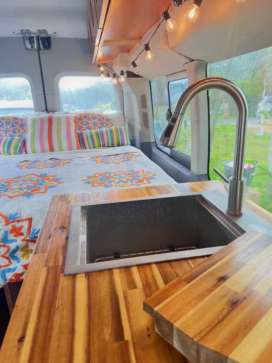 Cozy RV rental interior with a modern kitchen, wooden countertops, and a comfortable bed. String lights add warmth.. Ford Transit Custom 2016