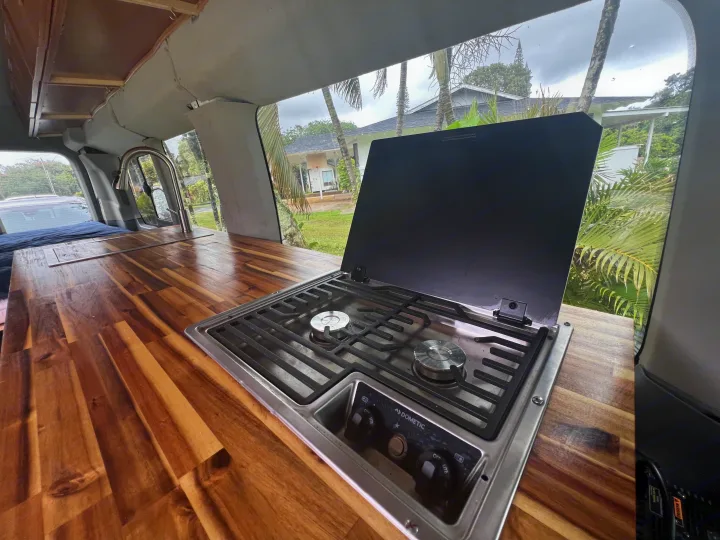 RV rental kitchen: wooden counter, Dometic two-burner stove with open lid, and a shiny faucet for cooking.. Ford Transit Custom 2016