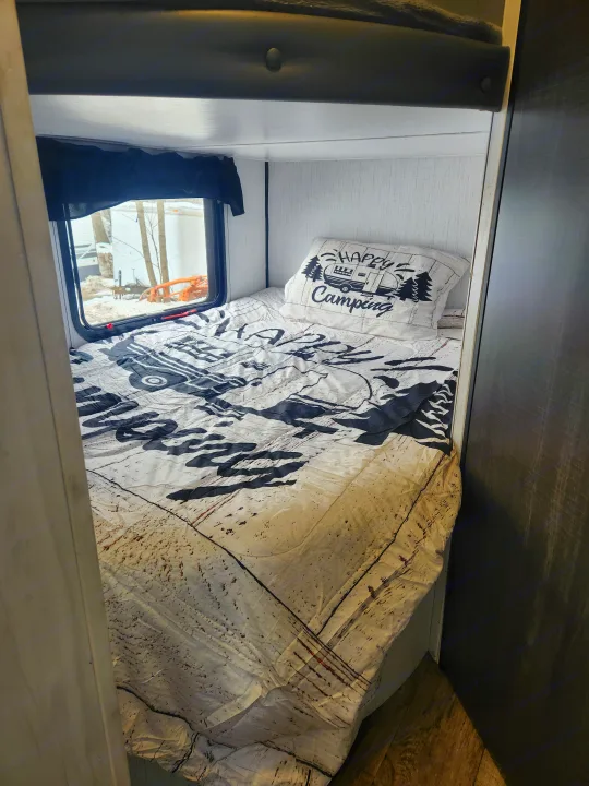 Cozy RV rental bunk bed with a 'Happy Camping' pillow, patterned bedding, and a window offering a snowy outdoor view.. Heartland RVs Trail Runner 2022