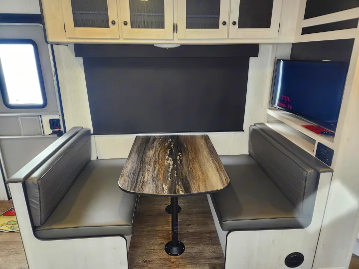 RV rental dinette: marbled table, gray benches, overhead cabinets, a window with a blackout shade, and a TCL Roku TV.. Heartland RVs Trail Runner 2022