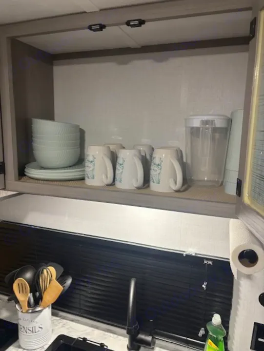 Inside an RV rental: a tidy kitchen with light blue dishes in an overhead cabinet, sink below, and utensils by the faucet.. Coleman Lantern 2024