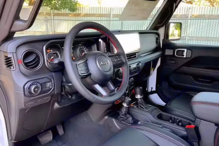 Inside a Jeep's driver cabin, featuring the black dashboard, steering wheel with red stitching, and central console controls. Ideal for an rv rental.. Jeep Wrangler Rubicon 2024