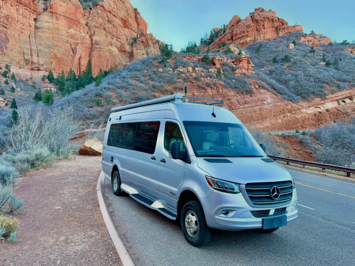 Silver Mercedes Sprinter RV rental parked by a scenic road, surrounded by towering red rock formations and desert plants.. Winnebago Era 2020