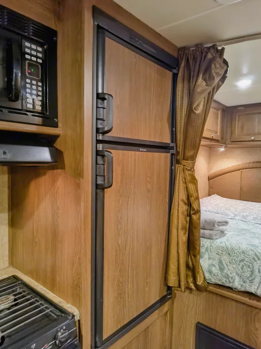 Inside an RV rental: a compact kitchen with microwave, stovetop, and Dometic fridge next to a bedroom with a bed and cozy curtain.. Coachmen Leprechaun 2015
