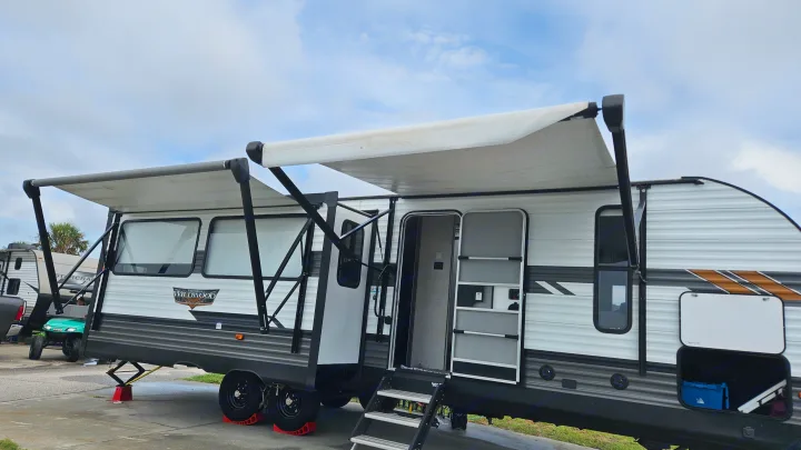 White/grey travel trailer RV rental with two extended awnings, an open screen door, and leveling jacks visible.. Forest River Wildwood 2021