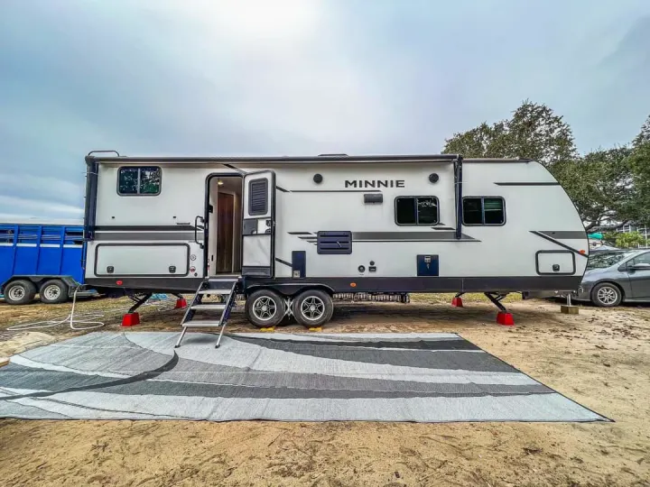 This spacious RV rental (Minnie travel trailer) is set up on a sandy campsite with an outdoor rug, ready for adventure.. Winnebago Minnie Winnie 2021