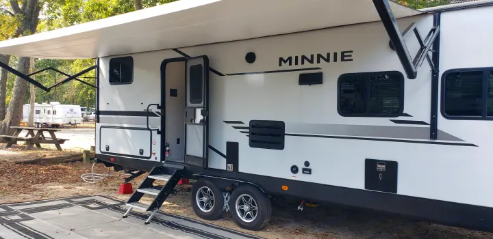 This white Minnie RV rental is set up at a leafy campground, with its awning out and steps down, ready for adventure.. Winnebago Minnie Winnie 2021