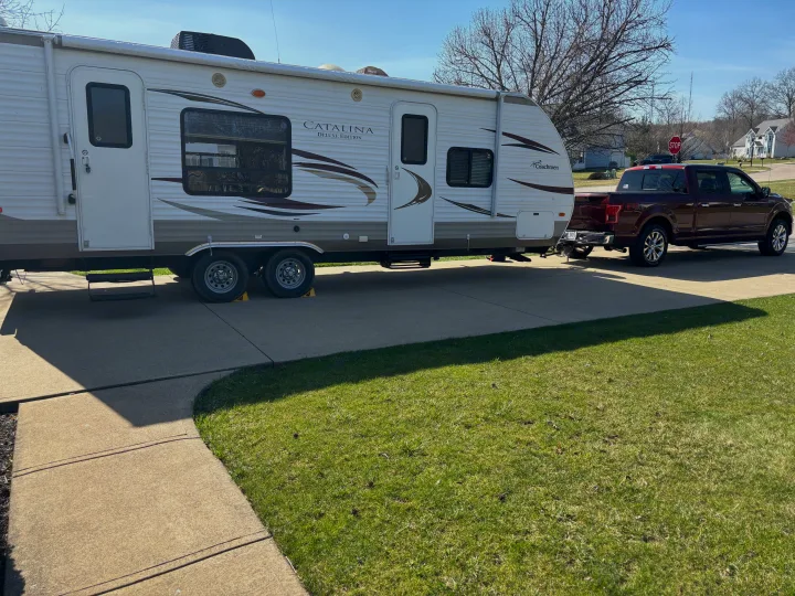 White Catalina RV rental hooked to a maroon Ford F-150 pickup truck on a sunny residential driveway, ready for a trip.. Coachmen Catalina Deluxe Edition 2013
