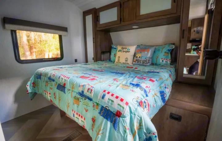 Experience comfort in this RV rental bedroom. Features a blue camper-themed bed, decor pillows, cabinets & window with nature views.. Forest River Salem 2022