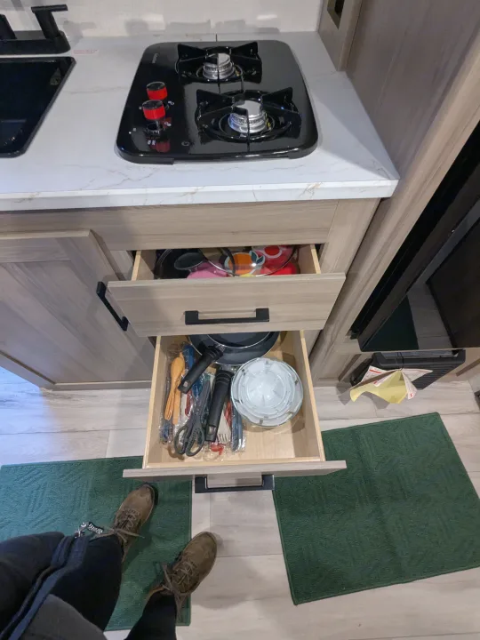 Compact RV rental kitchen featuring a two-burner stovetop and open drawers displaying organized cookware, bowls, & utensils.. Keystone Coleman 2025