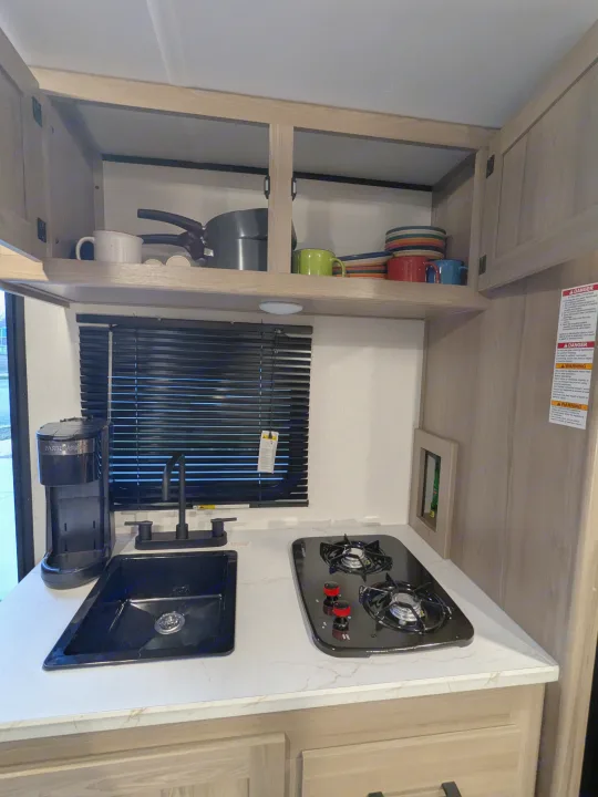 Compact RV rental kitchen with sink, two-burner stove, coffee maker, and overhead storage for dishes.. Keystone Coleman 2025