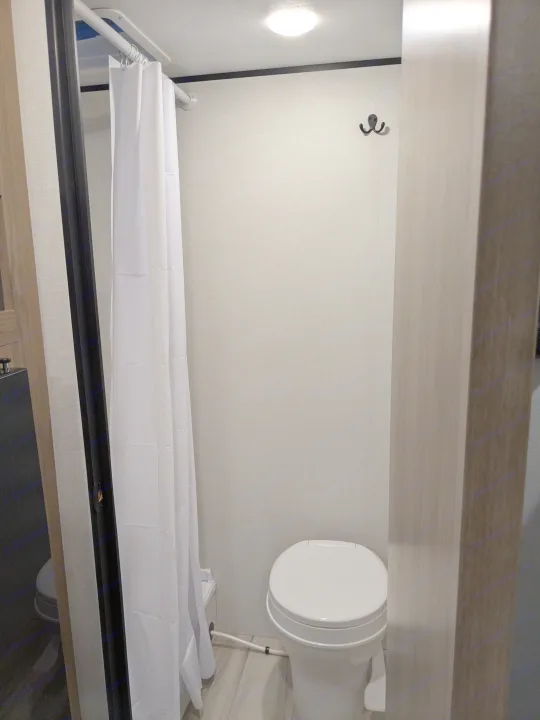 This RV rental bathroom features a compact design with a white toilet, shower curtain, overhead light, and wall hook.. Keystone Coleman 2025