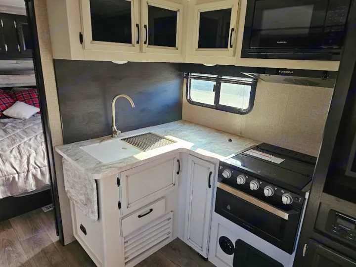 This RV rental shows a light-colored kitchen with a sink, black stove, microwave, white cabinets, and a bed in the background.. Jayco Jay Feather 2021