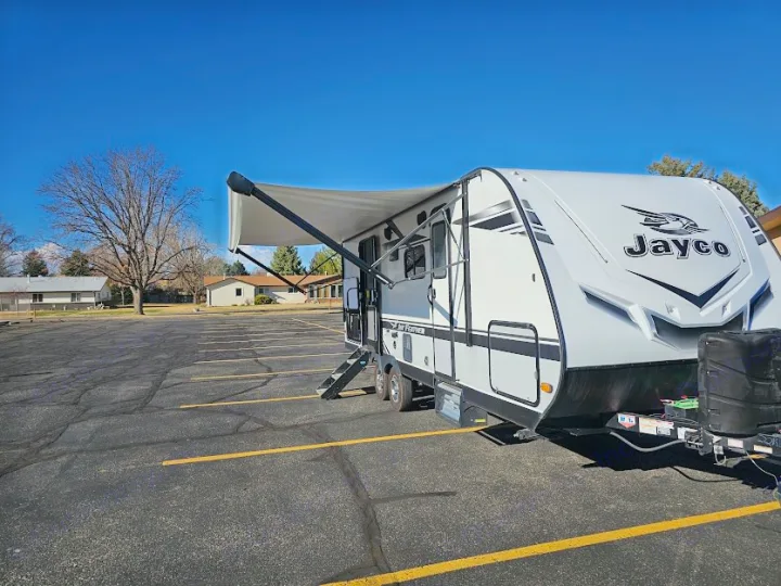 A white Jayco travel trailer, awning extended, awaits your next RV rental adventure in a sunny asphalt lot.. Jayco Jay Feather 2021