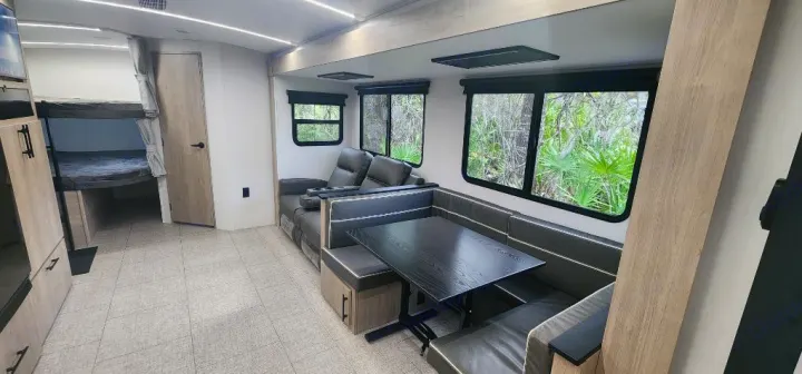 Bright RV rental interior showing bunk beds, a dinette, lounge seating, and windows with a view of lush greenery.. Grand Design Imagine 2800BH 2025