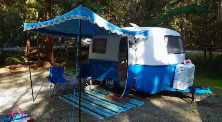 A blue and white vintage camper trailer with an awning and chairs at a shaded forest campsite. Explore RV rental options!. Boler Boler 1974