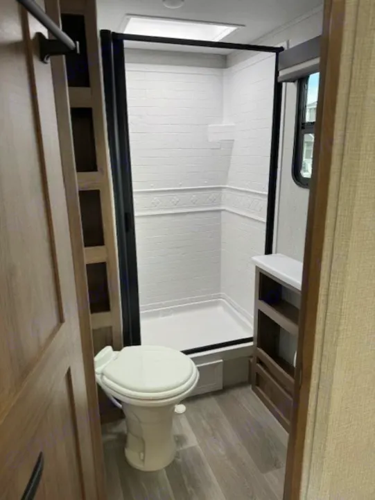 Inside an RV rental bathroom, featuring a white toilet, shower stall with clear door, and shelving. Light-colored interior.. Grand Design Imagine 2024