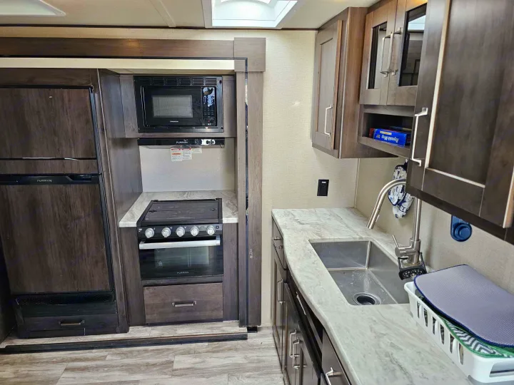RV rental kitchen: dark wood-look fridge, microwave, oven, and a light-marbled counter with a stainless steel sink.. Grand Design Transcend Xplor 2022