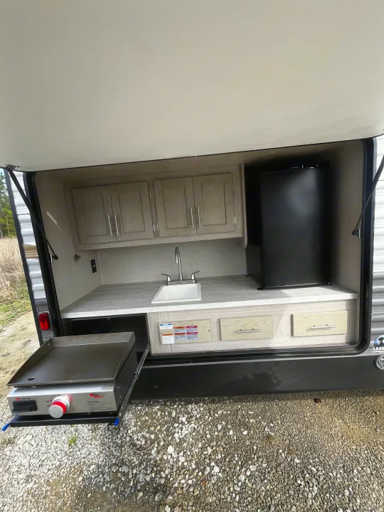 Outdoor kitchen setup on an RV rental, showing a sink, compact fridge, cabinets, and a pull-out griddle. Ideal for camping.. Forest River Aurora 2022