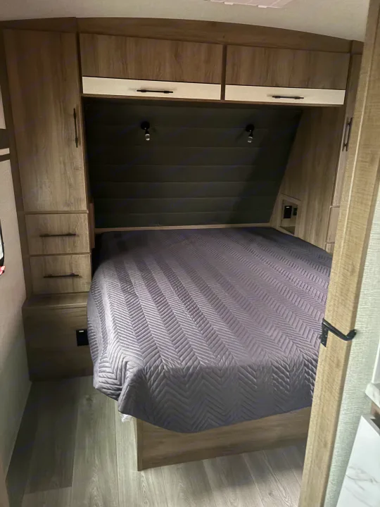 RV rental bedroom: A comfortable gray quilted bed flanked by wooden cabinets, reading lights, and power outlets.. Grand Design Imagine 2800bh 2025