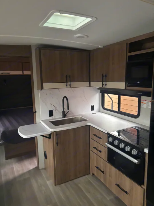 RV rental interior: modern kitchen with wood-grain cabinets, marble-look counters, sink, stove, and microwave.. Grand Design Imagine 2800bh 2025