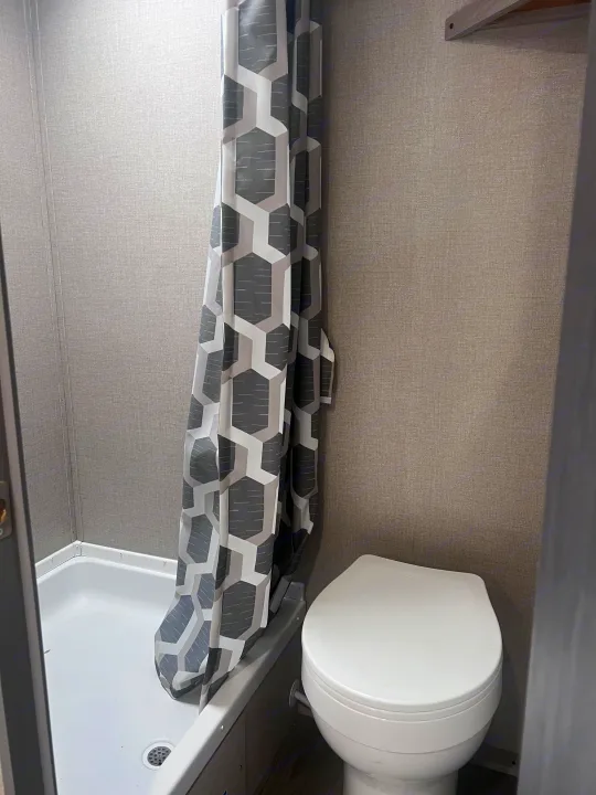 Inside an RV rental: a compact bathroom featuring a patterned gray and white shower curtain, white shower stall, and toilet.. Coachmen 164BHX 2023