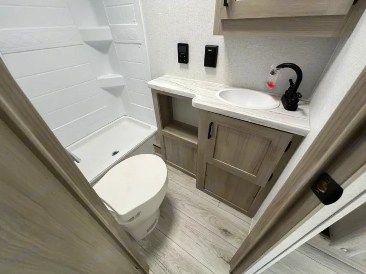 This RV rental features a modern, compact bathroom with a white shower, toilet, and sink vanity. Perfect for travel.. East to West Della Terra 181BH 2025