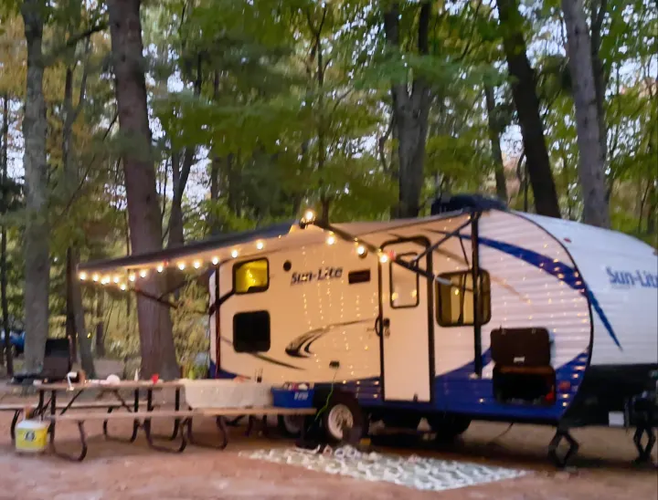 Cozy Sun-Lite rv rental at dusk, illuminated with string lights, perfect for a relaxing campsite evening with a picnic table.. Sunset Park RV Sun Lite 2019