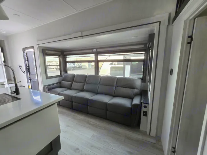 Inside a modern RV rental, featuring a spacious living room with a grey reclining sofa, large windows, and a kitchen counter.. Forest River East to West Ahara 390DS 2025