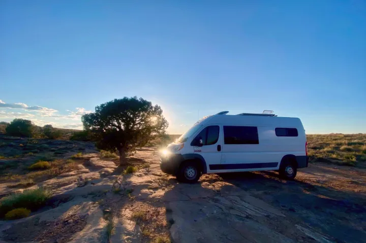 White camper van boondocking on rocky desert landscape at sunset, with sunrays peeking through. Your RV rental adventure awaits!. RAM Promaster 2500 159wb 2018