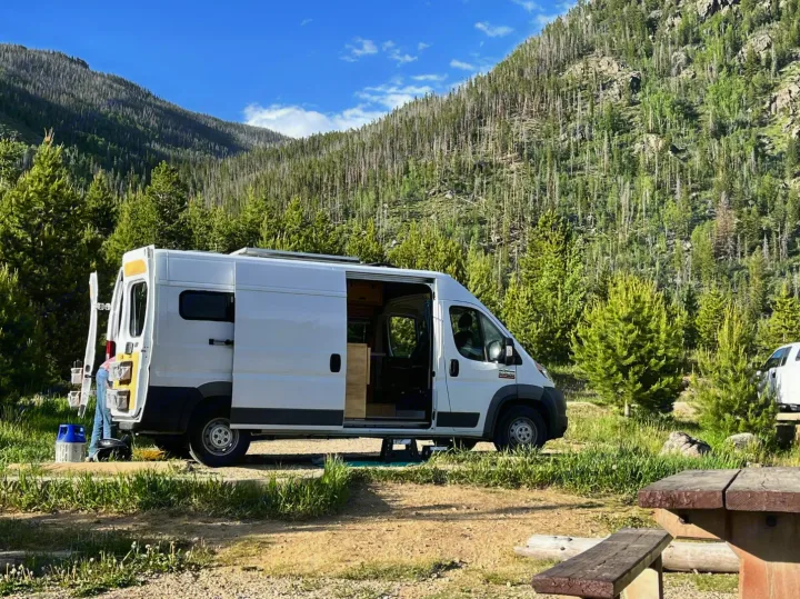 A white RV rental camper van is parked with its side door open at a scenic mountain campsite surrounded by green and bare trees.. RAM Promaster 2500 159wb 2018