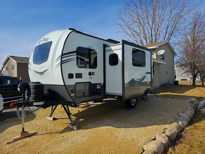 A white & gray travel trailer, with its slide-out extended, makes a great RV rental. Parked on a gravel lot beside a house.. Forest River Flagstaff E-Pro 2024
