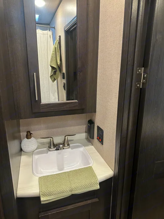 RV rental bathroom with a dark wood sink cabinet, mirror, and wall-mounted controls. A white sink and green towel are visible.. Forest River X-Lite 2017