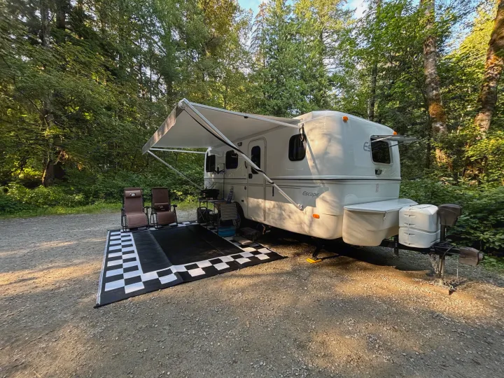 This RV rental is set up for a cozy camping experience with an extended awning, chairs, and mat on a gravel site surrounded by trees.. Escape Trailer Escape Trailer Trailer 2022