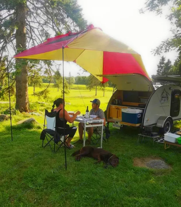A couple and dog enjoy an outdoor meal by their RV rental teardrop trailer with open kitchen setup.. Car-gotrailers liberty2goXL 2021