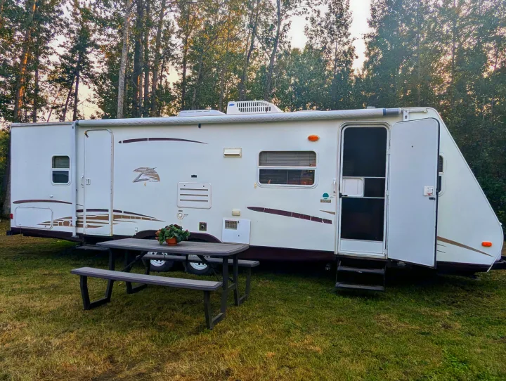 White travel trailer RV rental with open door & steps, parked next to a picnic table on green grass, surrounded by trees.. Keystone RV Zeppelin Z-II 2006