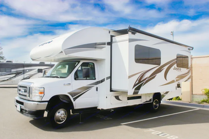 A white Jayco Redhawk Class C motorhome, an RV rental, shows its extended side slide-out in a sunny parking lot.. Jayco Redhawk 2022