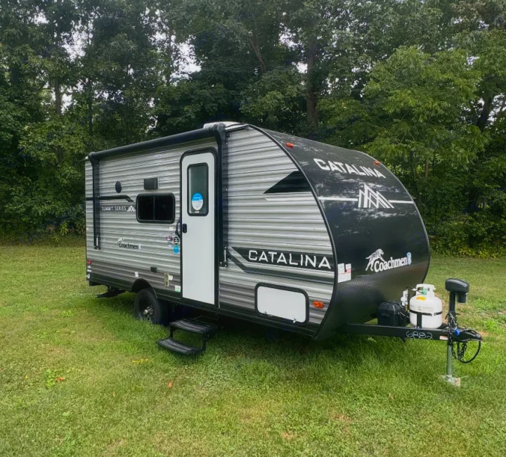This black & gray Catalina travel trailer, ideal for your next RV rental, sits on grass amidst trees, ready for adventure.. Forest River Coachmen Catalina Summit 2024
