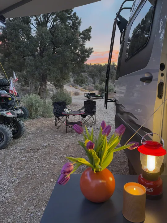 Relaxing at an RV rental campsite at sunset. Features cozy seating, floral decor, a lantern, and an ATV.. Winnebago Revel 2023