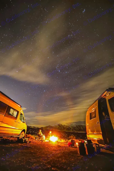 RV rental adventure: Two camper vans, people by a campfire, & a stunning starry night sky over mountains.. Winnebago Revel 2023