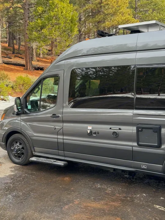 Grey rv rental camper van parked in a lush forest. It's a dark grey camper with roof solar panels, side steps, and utility hookups, reflecting the trees in its windows on a damp road.. Thor Sanctuary 2023