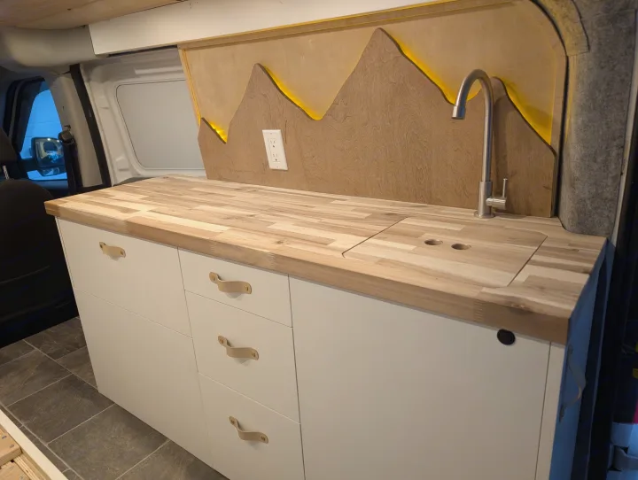 RV rental interior: custom kitchen featuring a wood block countertop, covered sink, and backlit mountain wall panel design.. Dodge Sprinter Van 2015