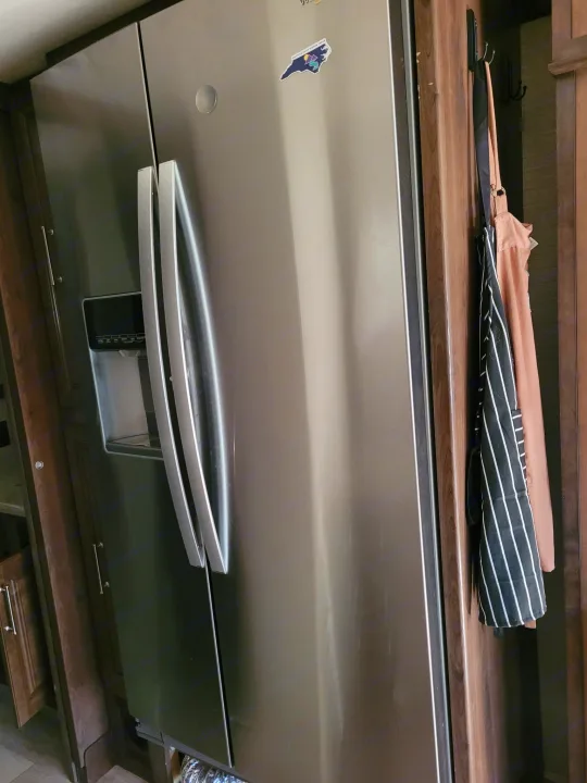 Modern stainless steel side-by-side fridge with water dispenser in an RV rental kitchen. Hooks with apron visible. Entegra Accolade 2020