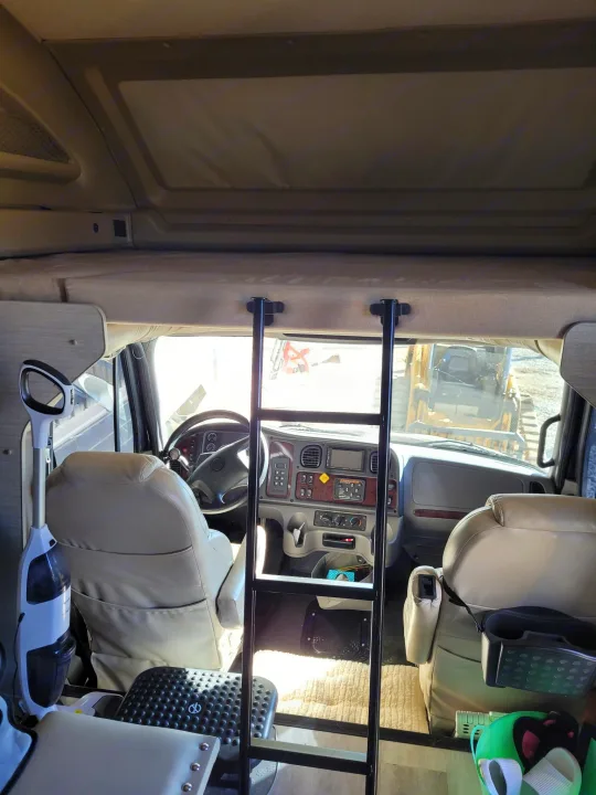 RV rental interior: View into the cockpit with tan seats, steering wheel, and dashboard. Overhead bunk accessible via a black ladder.. Entegra Accolade 2020