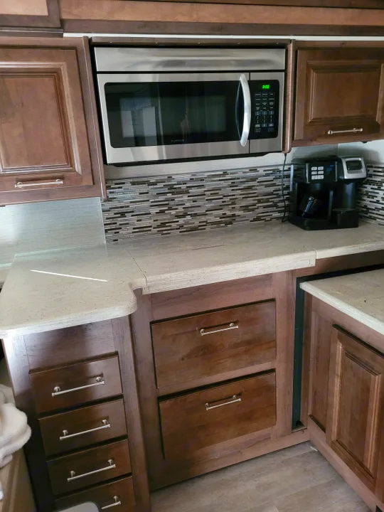 This RV rental kitchen features a stainless microwave, wood cabinets, and light speckled counters with a coffee maker.. Entegra Accolade 2020