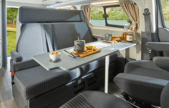 This rv rental campervan's cozy interior features a dining table with breakfast items, overlooking a scenic lake.. Ford Nugget 2025
