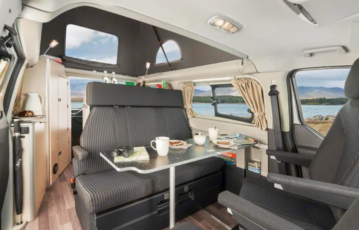 Inside an RV rental: a dinette set for two, framed by large windows revealing a scenic lake and mountain view, with a pop-top roof.. Ford Nugget 2025