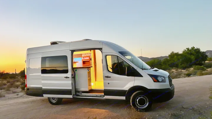 This white camper van RV rental, with its inviting, lit interior, is parked on a desert road under a beautiful sunset.. Ford Transit 2019