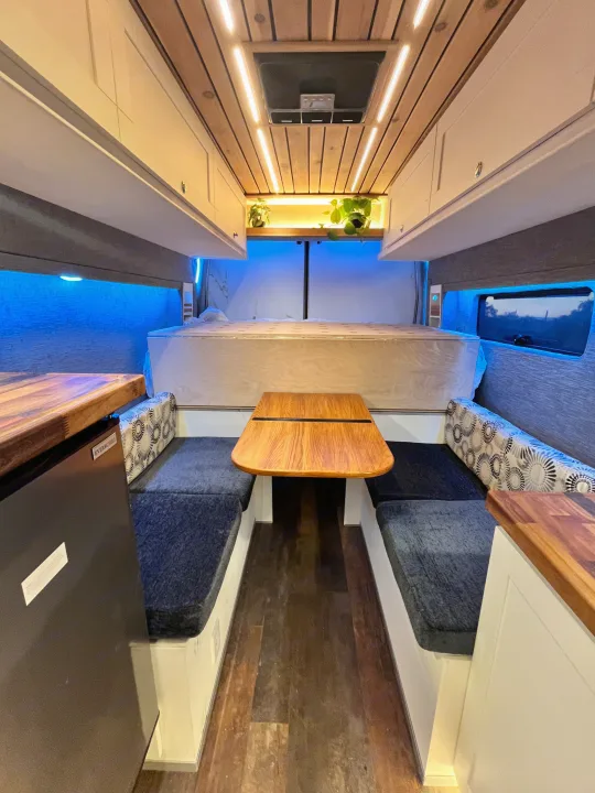 An RV rental's cozy interior: dining table, benches, bed, kitchen counter, and ample storage. Ambient blue lighting.. Ford Transit 2019